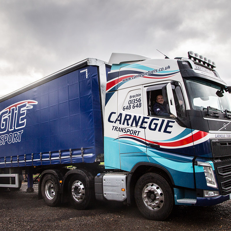 A man smiling out the window of the driver's seat of a Carnegie Transport road haulage lorry with the window down.